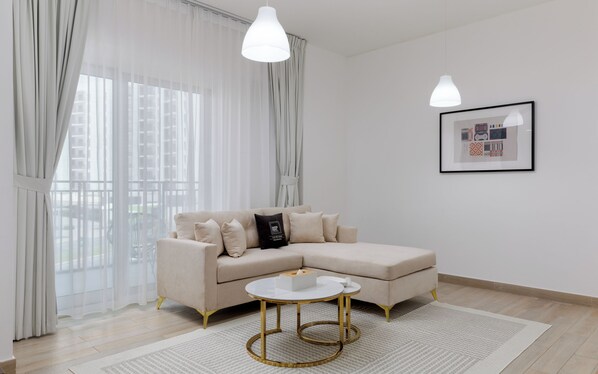 A modern living room with a beige sectional sofa, a round coffee table with gold legs, sheer curtains, and a framed picture on the wall.