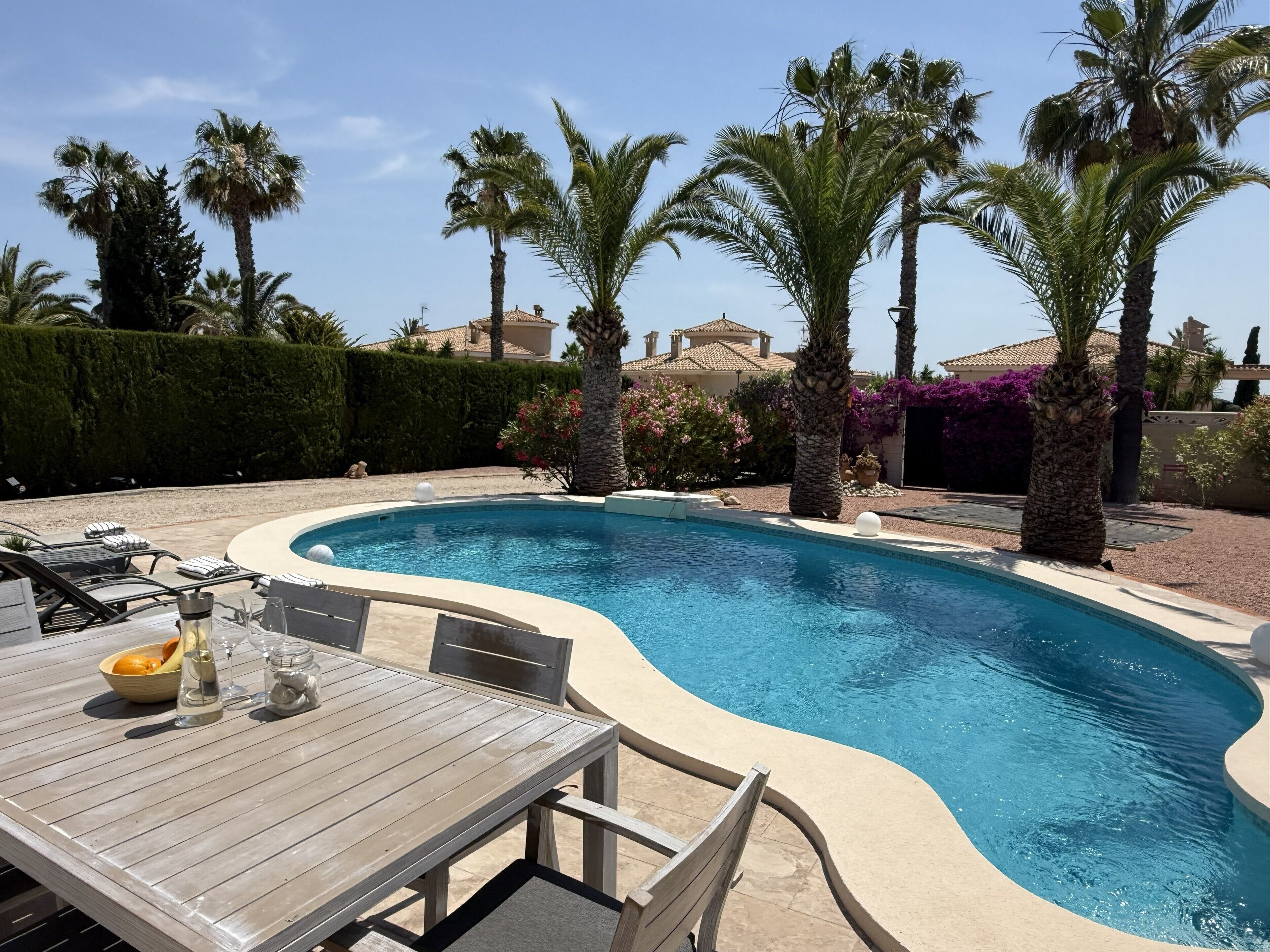 Une piscine à bord incurvé, entourée de palmiers et dotée d’une terrasse en bois avec des meubles d’extérieur.