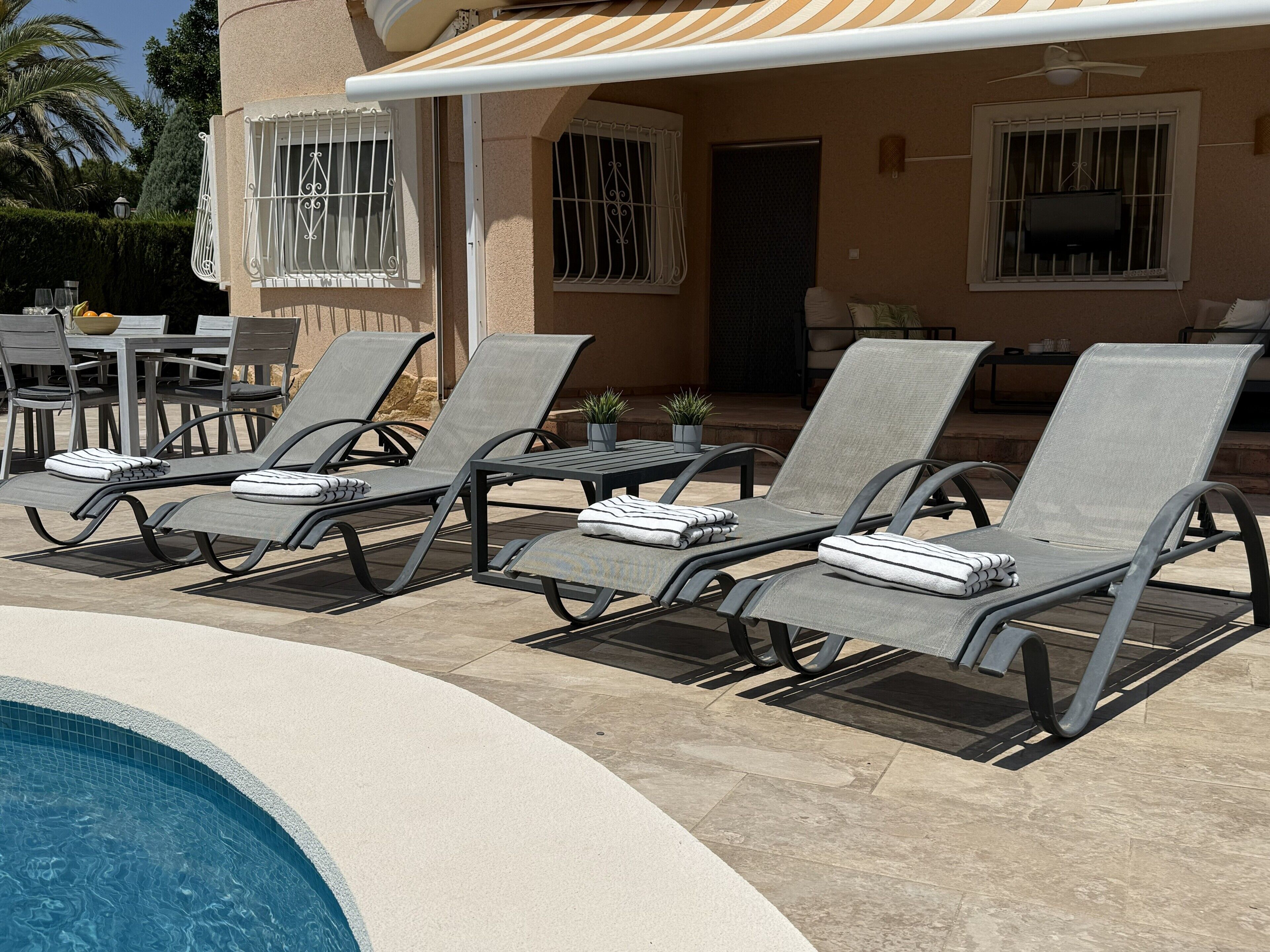 Un espace aménagé au bord de la piscine, avec des chaises longues et une table, une terrasse couverte et un bâtiment dont les fenêtres sont protégées par des barreaux.