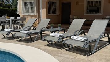 Un espace aménagé au bord de la piscine, avec des chaises longues et une table, une terrasse couverte et un bâtiment dont les fenêtres sont protégées par des barreaux.
