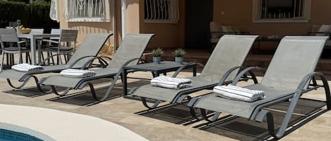 A poolside area with lounge chairs and a table, a covered patio, and a building with barred windows.