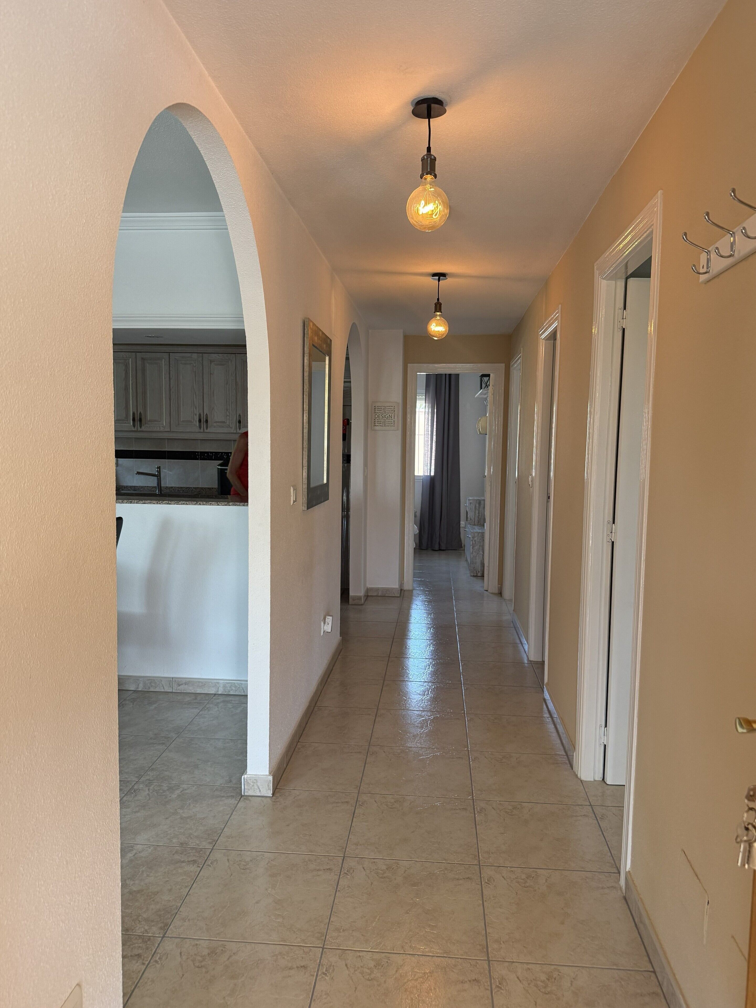 A hallway with a light fixture, a door with a coat rack, and a kitchen area with cabinets.
