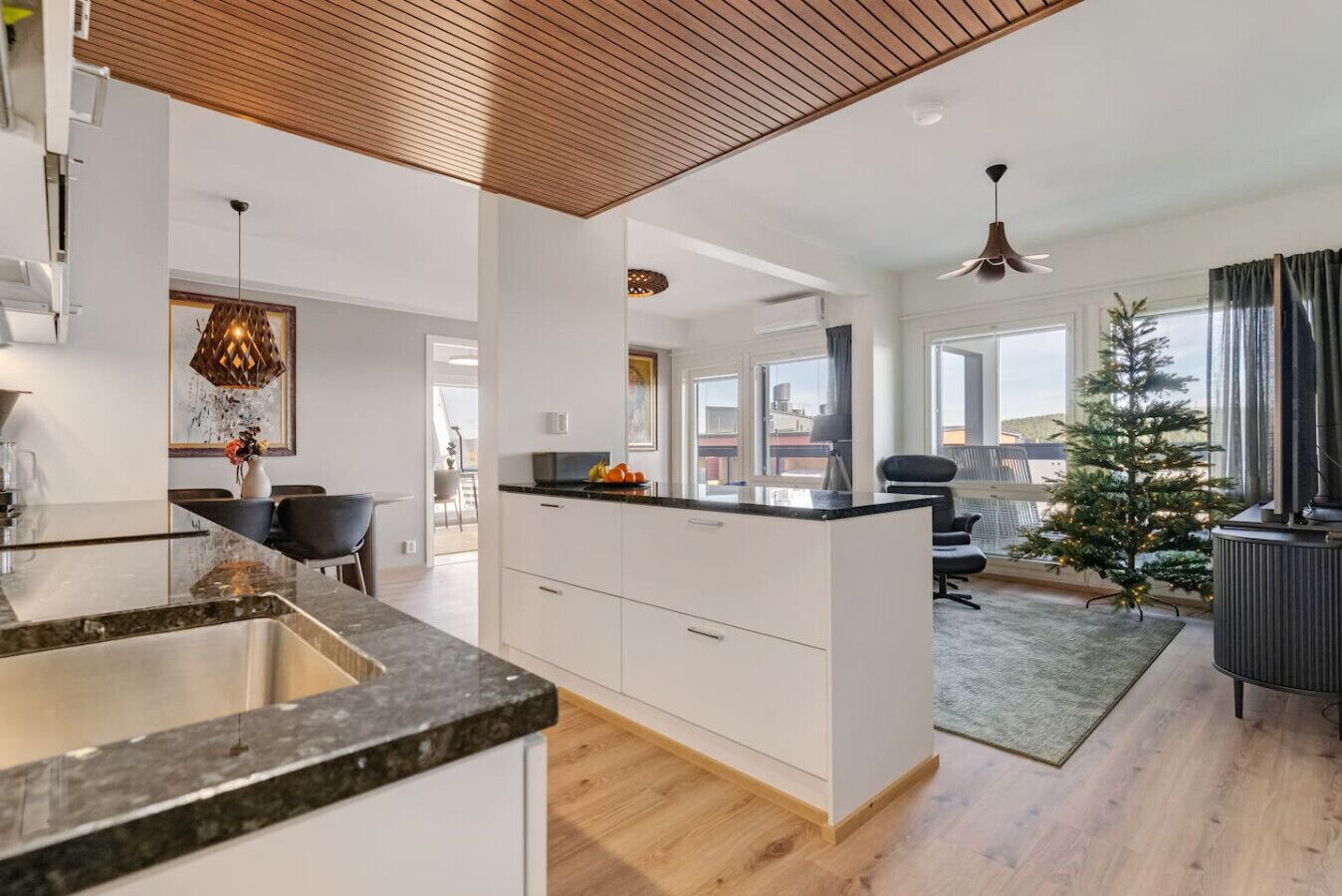 A modern kitchen with a central island, a dining area with chairs, a small Christmas tree, and a view of the city through large windows.