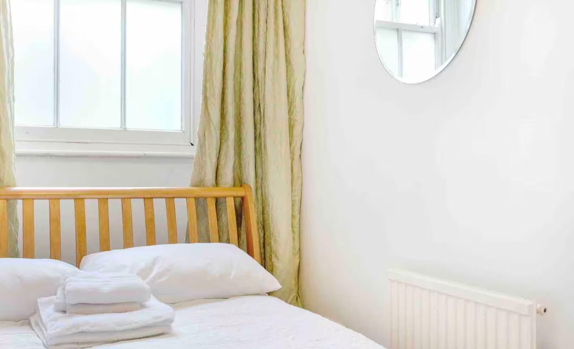 Ein Schlafzimmer mit Holz-Kopfteil, einem Bett mit weißer Bettwäsche, einem Heizkörper, einem Fenster mit Vorhängen und einem runden Spiegel an der Wand.