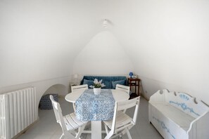 A small, white, attic-style room with a round table, white chairs, a blue-patterned tablecloth, a white bench, and a small table with a lamp. - A cozy 48-square-meter apartment, ideal for two people, located on the first floor of a typical historic center home (accessible via stairs). The location is perfect: the property is right in the town center (within the ZTL/restricted traffic zone), with (Martina Franca)