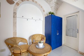 Une pièce avec une porte cintrée peinte en blanc, un meuble bleu, deux chaises en osier et une petite table sur laquelle est posée une plante.