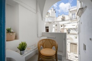 Un balcon étroit avec une chaise en osier, des plantes en pot et une vue sur les bâtiments voisins.