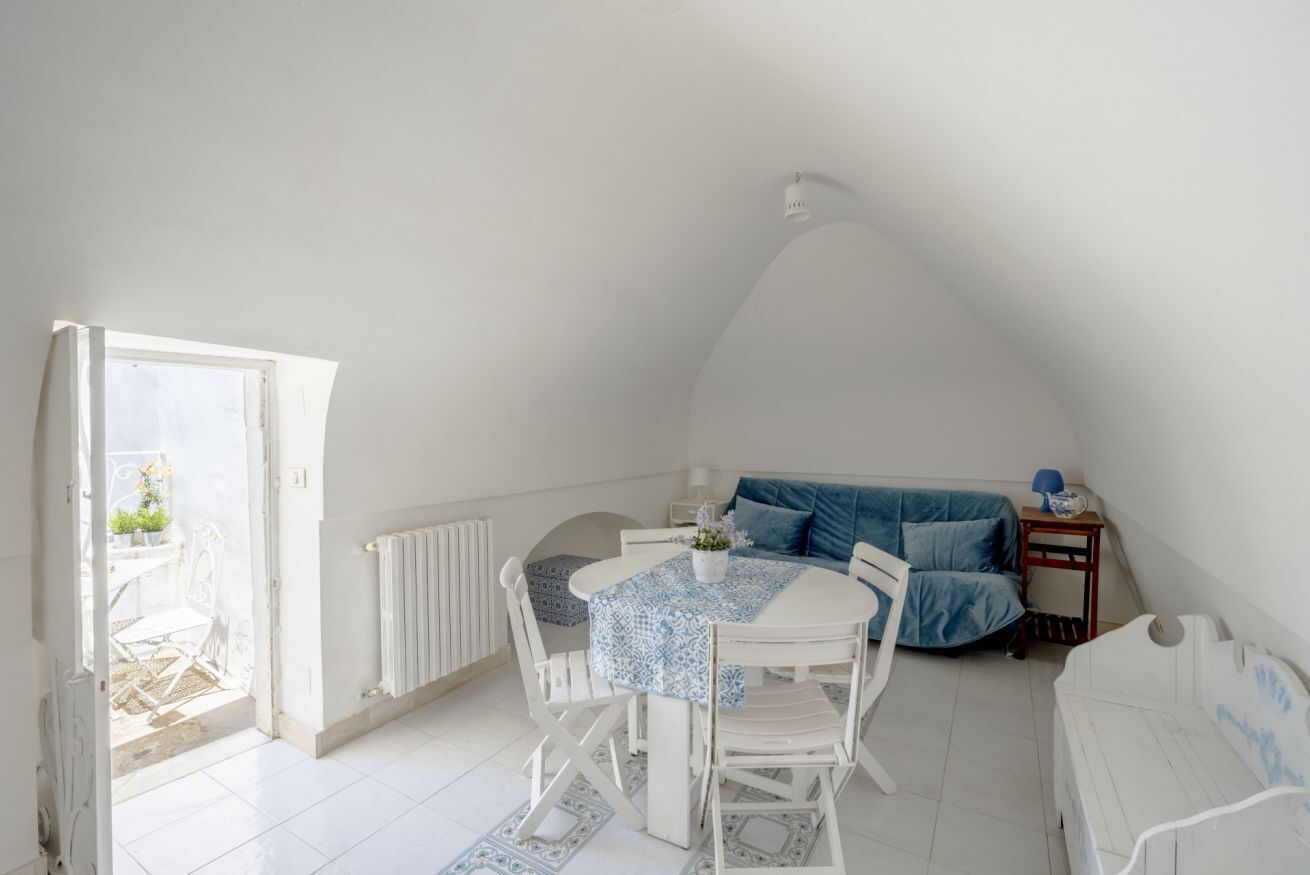 Una habitación con una mesa blanca, sillas, un sofá azul y una mesita de noche con una lámpara.