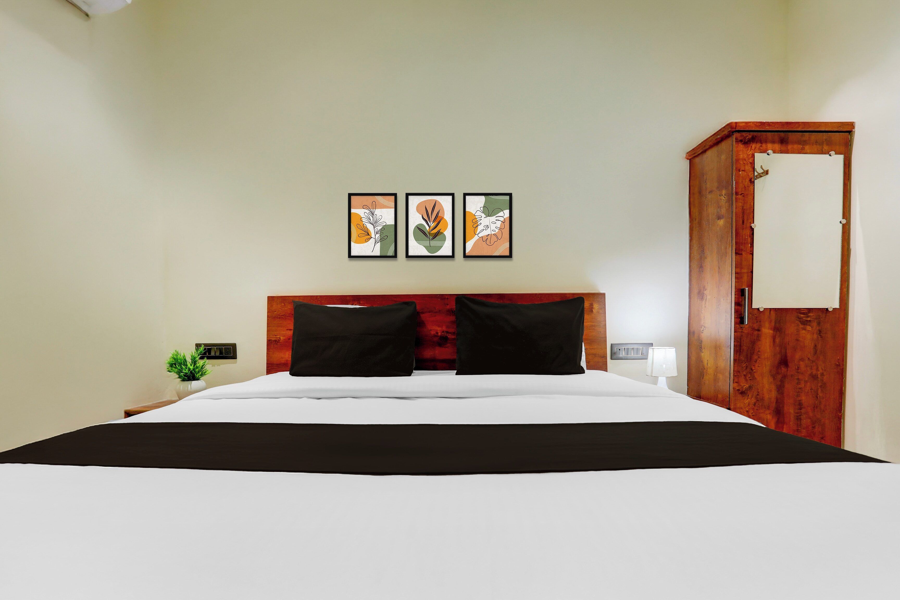 A hotel room with a bed, a nightstand, a lamp, a wooden wardrobe, and three framed artworks on the wall.