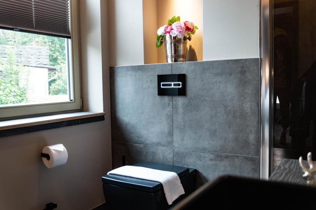 Un baño con inodoro, secamanos, dispensador de toallas de papel, un pequeño banco y un interruptor de luz en la pared.