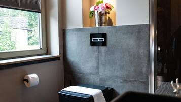 Un baño con inodoro, secamanos, dispensador de toallas de papel, un pequeño banco y un interruptor de luz en la pared.