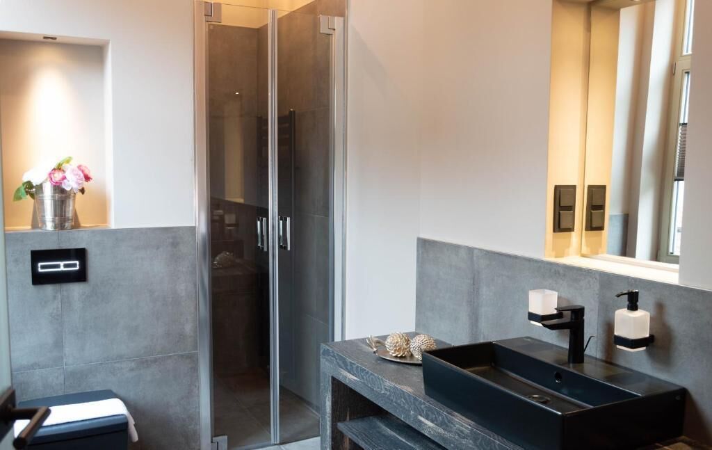 A modern bathroom with a glass shower enclosure, a dark sink with a soap dispenser, and a vase with flowers on the countertop.