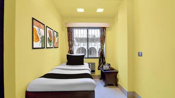 Une chambre d’hôtel étroite, avec un lit simple, une petite table sur laquelle se trouvent une bouilloire et des tasses, et une fenêtre avec des rideaux.