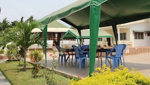 An outdoor seating area with blue plastic chairs and a covered table, surrounded by greenery and a building in the background. - Loreto Palm Villa: 17 en-suite AC rooms with on-site restaurant (Cape Coast, Ghana)