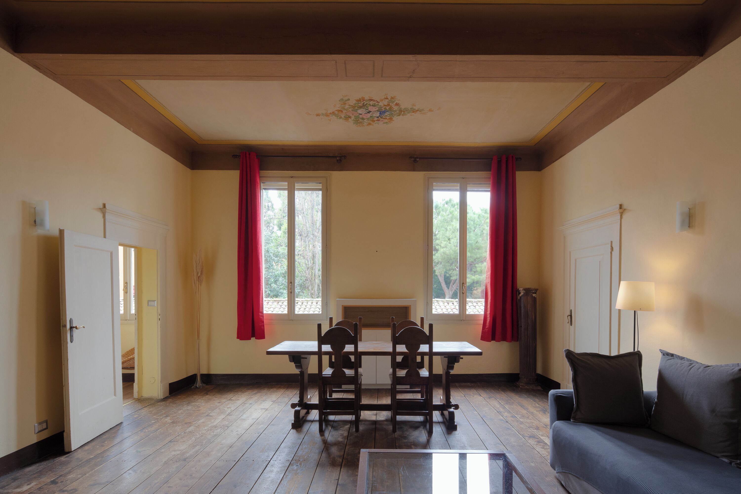 Ein Zimmer mit Holzboden, einem Tisch mit Stühlen, einem Sofa und zwei Fenstern mit roten Vorhängen.