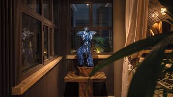 Eine Schaufensterpuppe in einem blauen Kleid steht auf einer Holzplattform, im Hintergrund sind Fenster und eine Pflanze zu sehen.