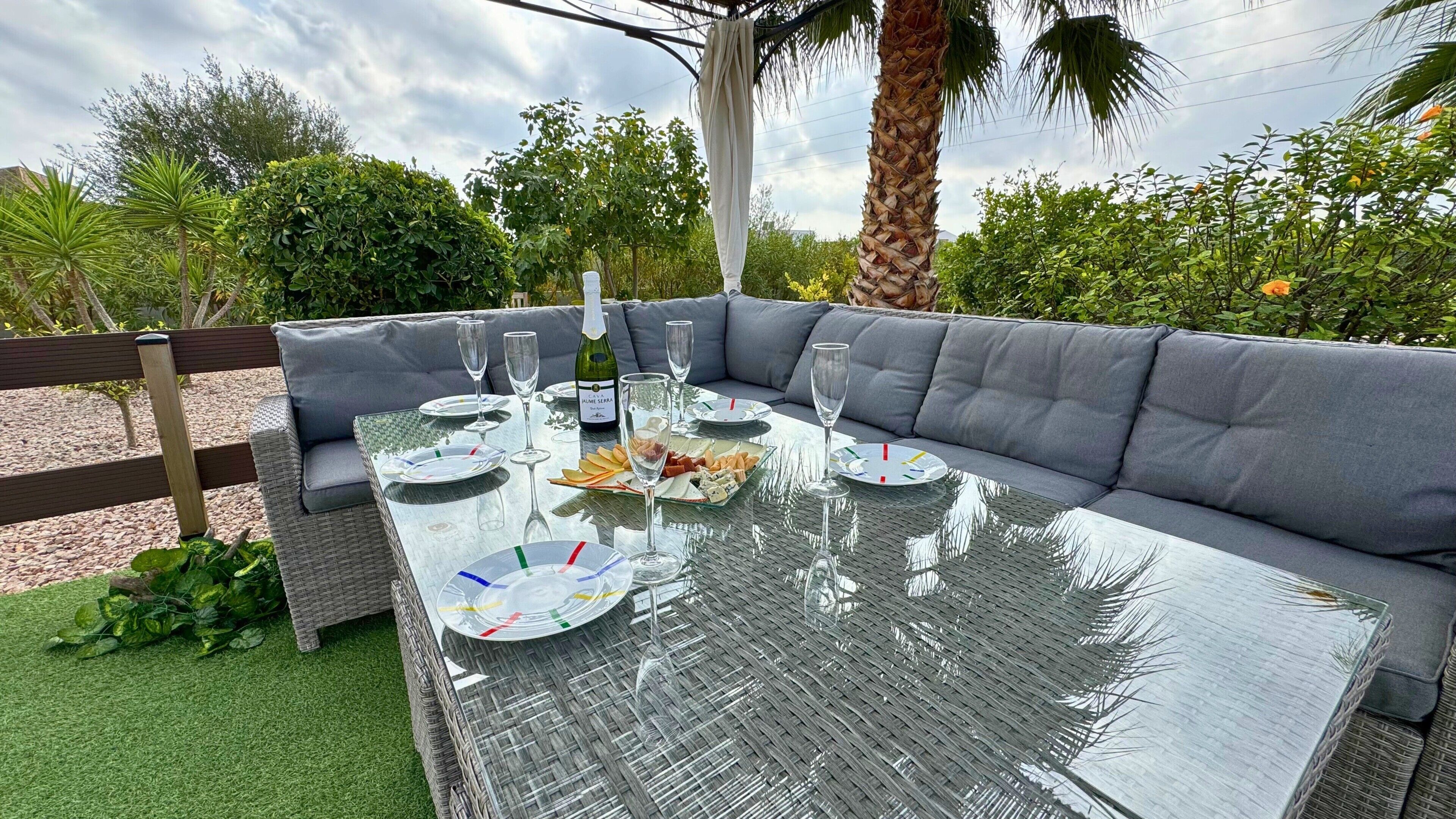 Un área de comedor al aire libre con una mesa de vidrio, asientos acolchados de color gris y una botella de vino.