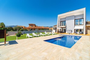 Un área de piscina moderna con una piscina azul cristalina, sillas de descanso blancas y un edificio de dos pisos con grandes ventanales.
