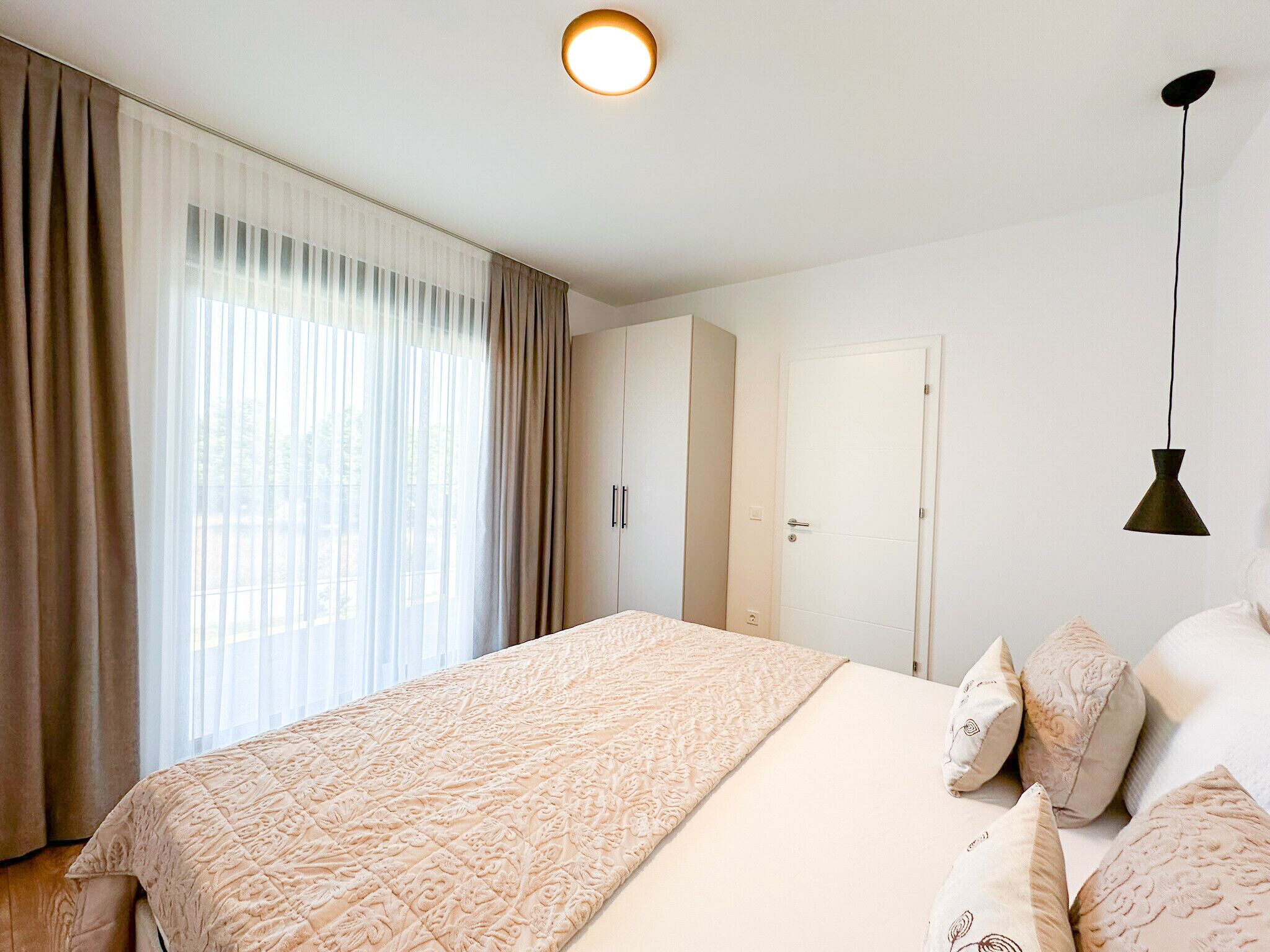 Un dormitorio con cama, cortinas, armario, puerta y una lámpara colgante.