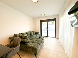 Ein modernes Wohnzimmer mit einem Ecksofa, einem kleinen runden Tisch und einem flach an der Wand befestigten Flachbildfernseher.