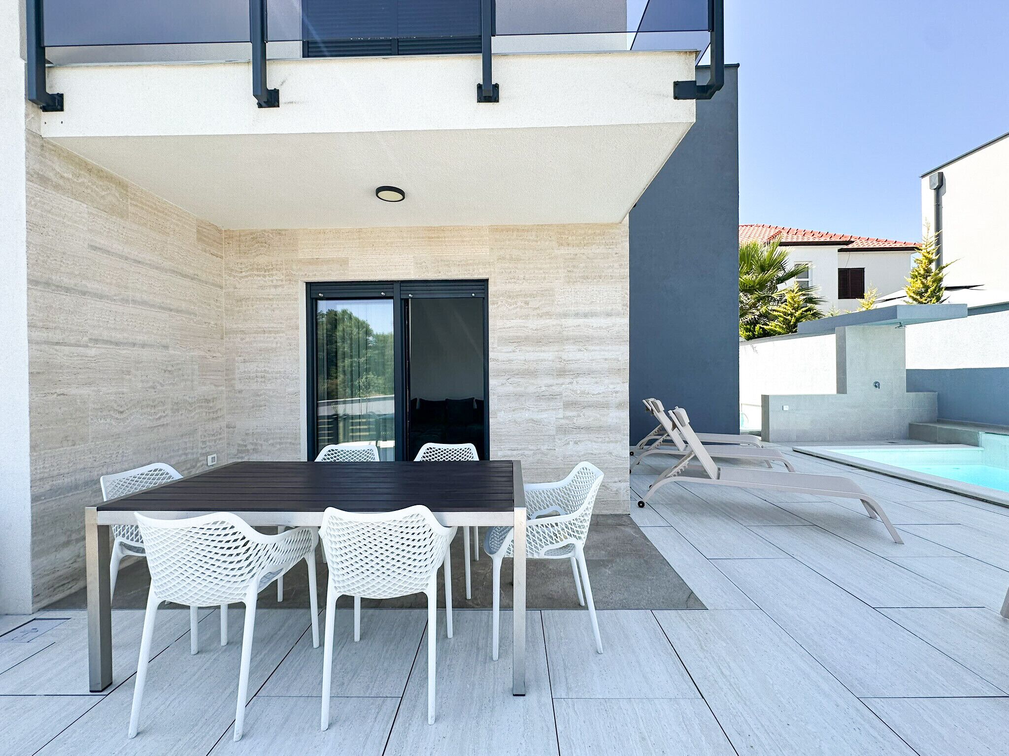Una zona d'àpat exterior moderna amb taula i cadires, una entrada amb porta de vidre i una zona de relaxació a la piscina.