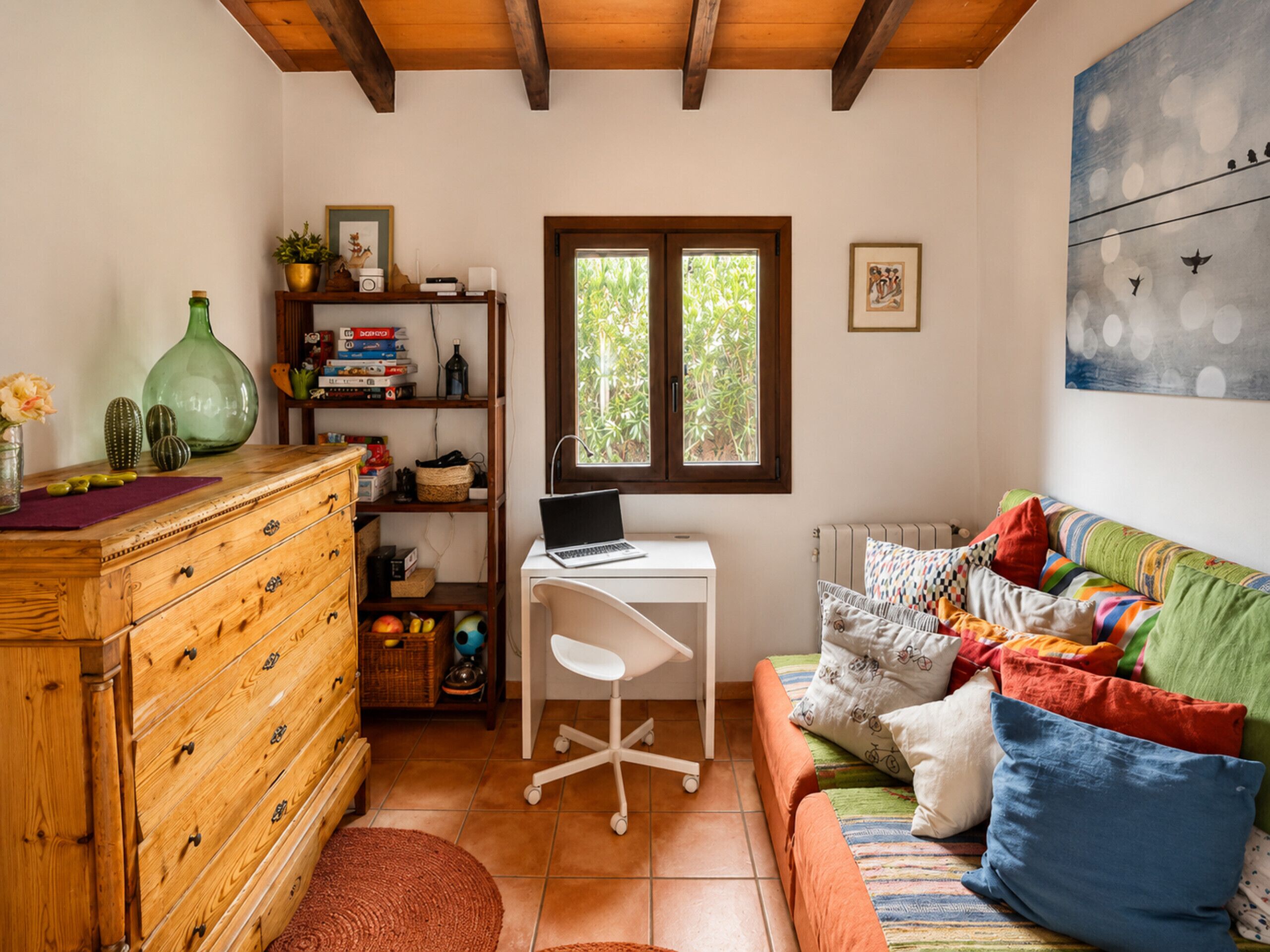 A cozy room with a wooden dresser, a desk with a laptop, a colorful sofa, and a window with a view of greenery.