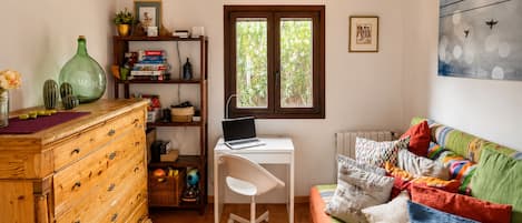A cozy room with a wooden dresser, a desk with a laptop, a colorful sofa, and a window with a view of greenery.