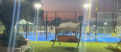 Una cancha de tenis exterior de noche con iluminación artificial, una zona de estar cubierta y un primer plano con césped.