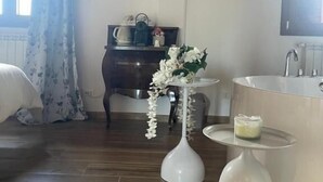 Una habitación con piso de madera, un sofá blanco, una mesa blanca con un florero, una mesita blanca con una vela, un televisor de pantalla plana montado en la pared y una bañera blanca grande.