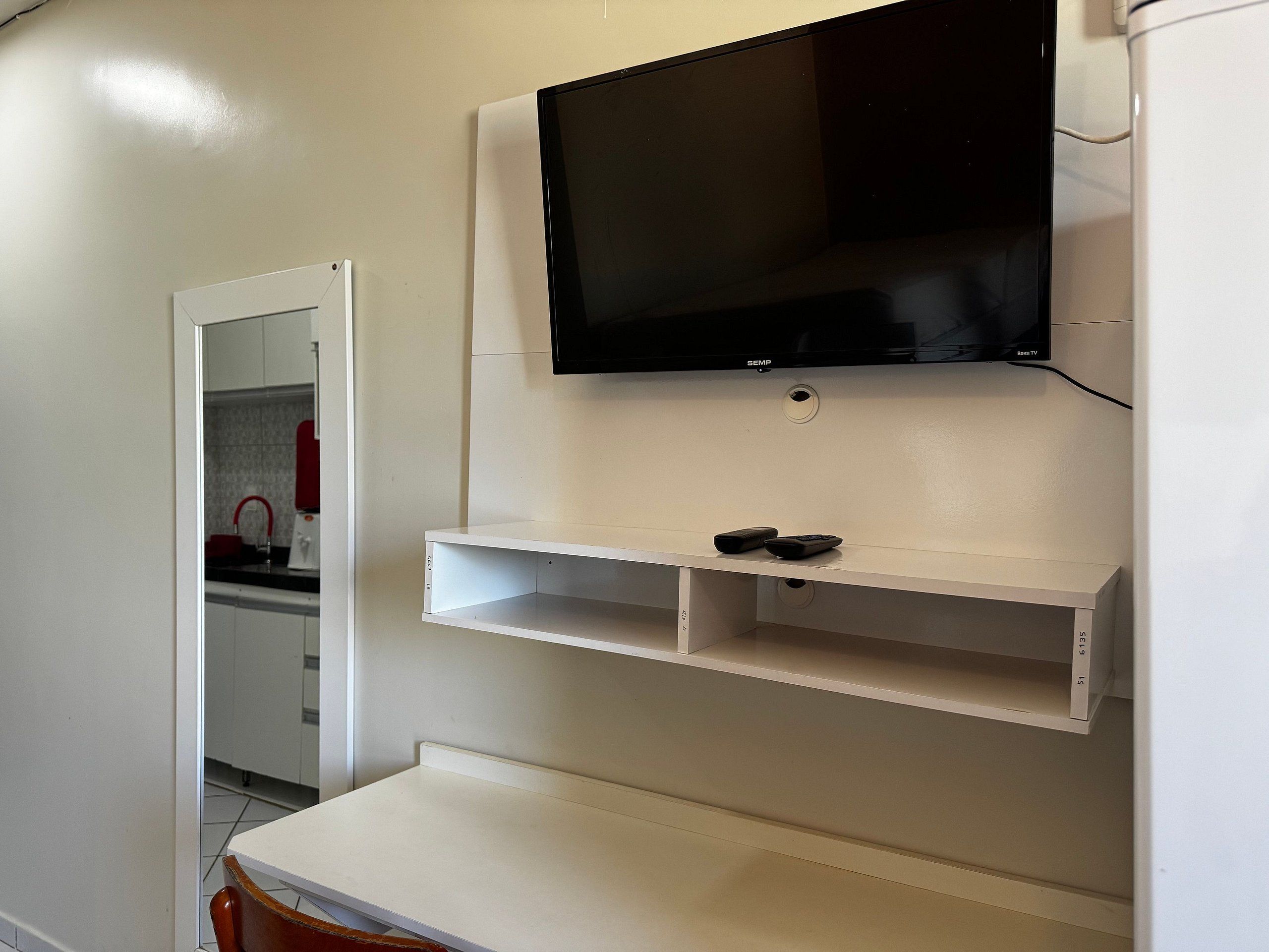 Quarto com televisão na parede, dois controles remotos em uma prateleira, uma mesa branca e uma área de cozinha com um eletrodoméstico vermelho.