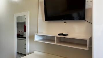 Quarto com televisão na parede, dois controles remotos em uma prateleira, uma mesa branca e uma área de cozinha com um eletrodoméstico vermelho.