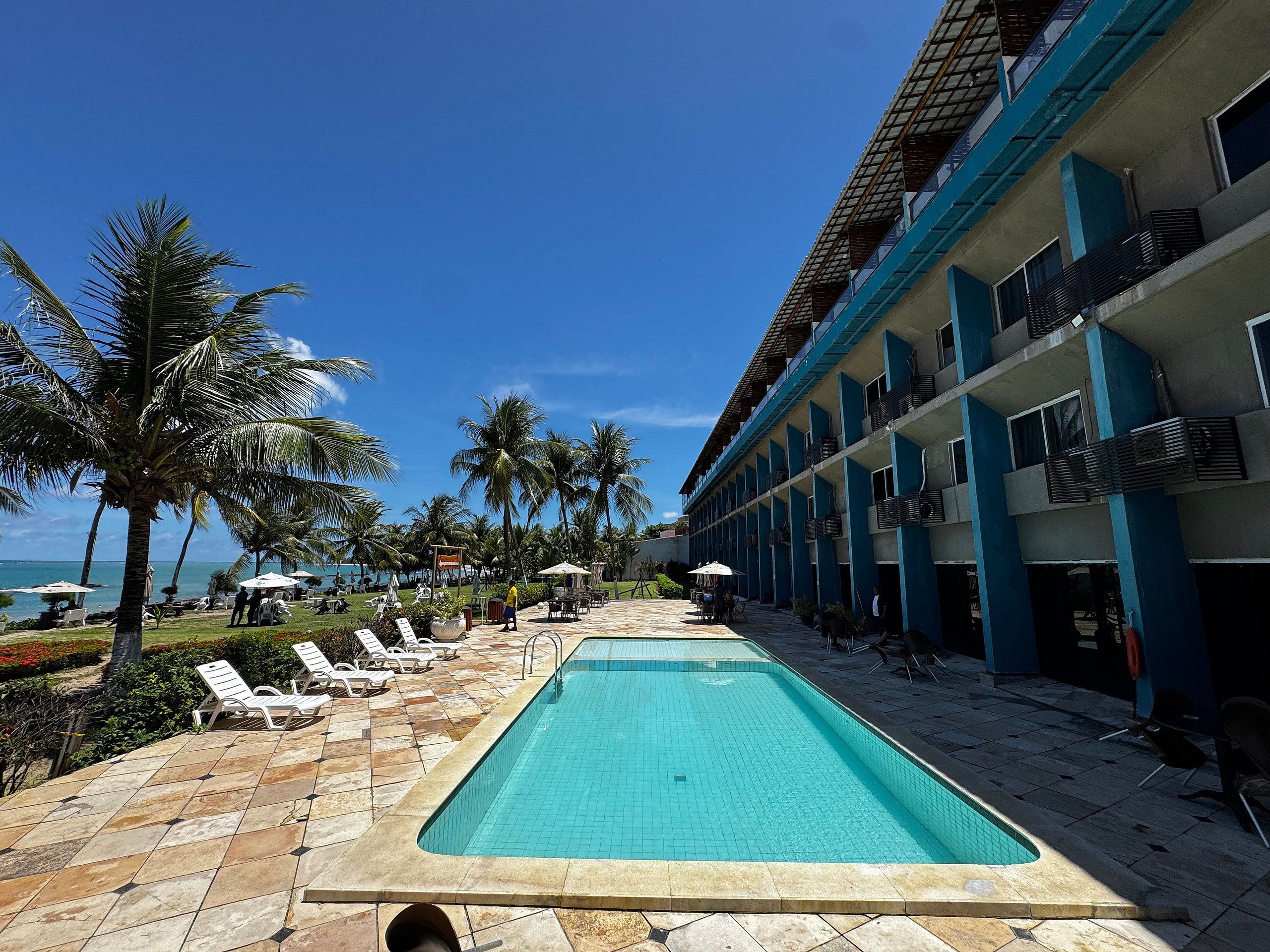Uma piscina com espreguiçadeiras e guarda-sóis, um edifício com varandas e palmeiras.