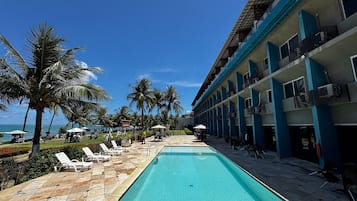 Uma piscina com espreguiçadeiras e guarda-sóis, um edifício com varandas e palmeiras.