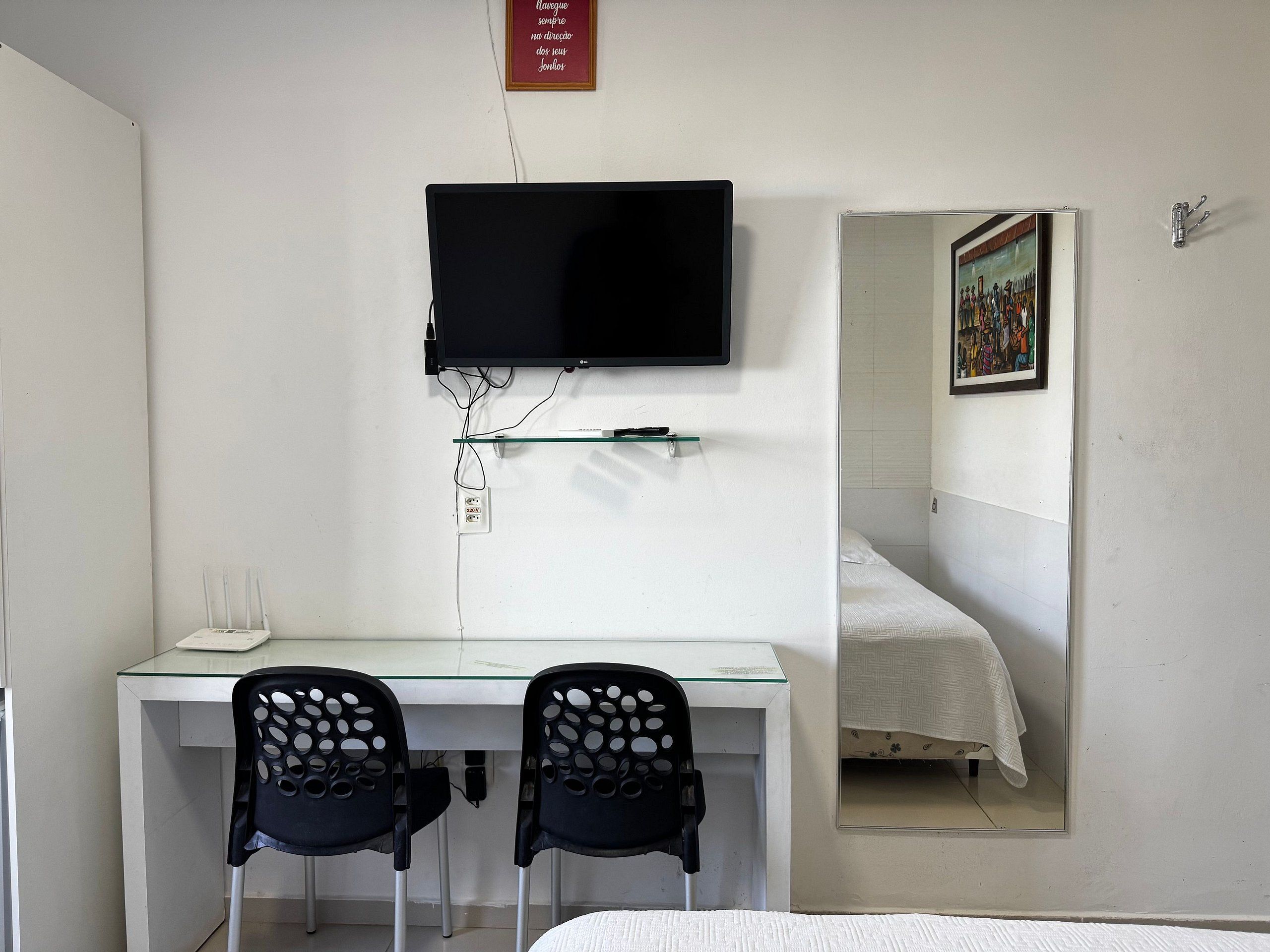 Un dormitorio con cama, escritorio con dos sillas, televisor montado en la pared y un espejo que refleja la habitación.