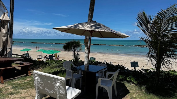 Een eetgedeelte aan het strand met een tafel, stoelen, een grote parasol en uitzicht op zee.