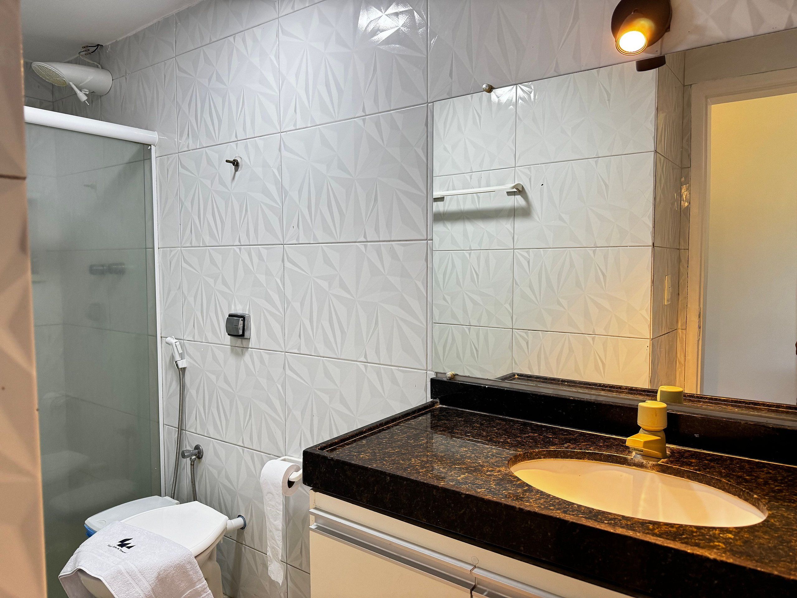 Un baño con cabina de ducha de vidrio, inodoro blanco, lavamanos de mármol con grifo amarillo y espejo con luz superior.