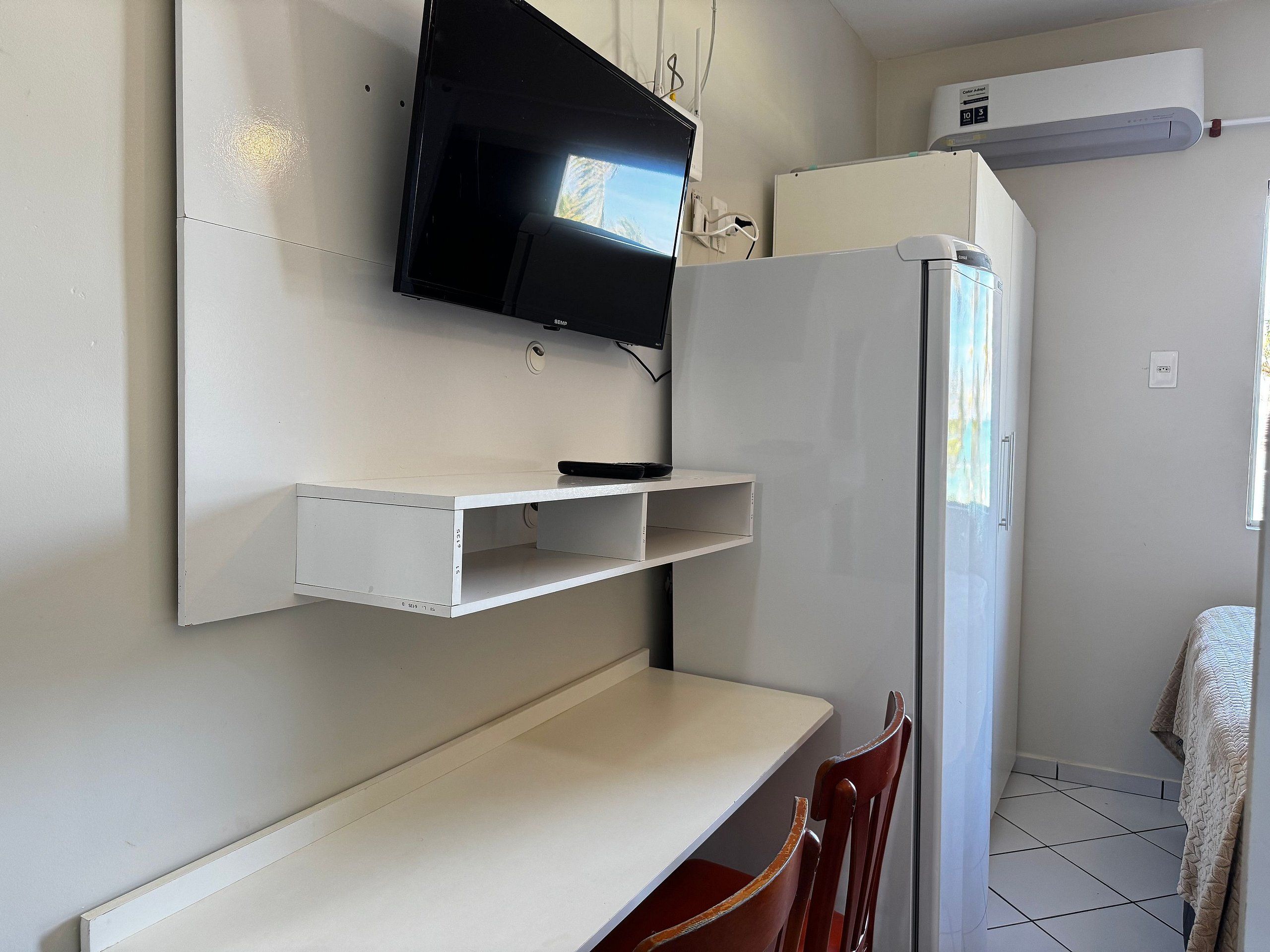 A room with a mounted television, a white shelf, a white table, and a white refrigerator.