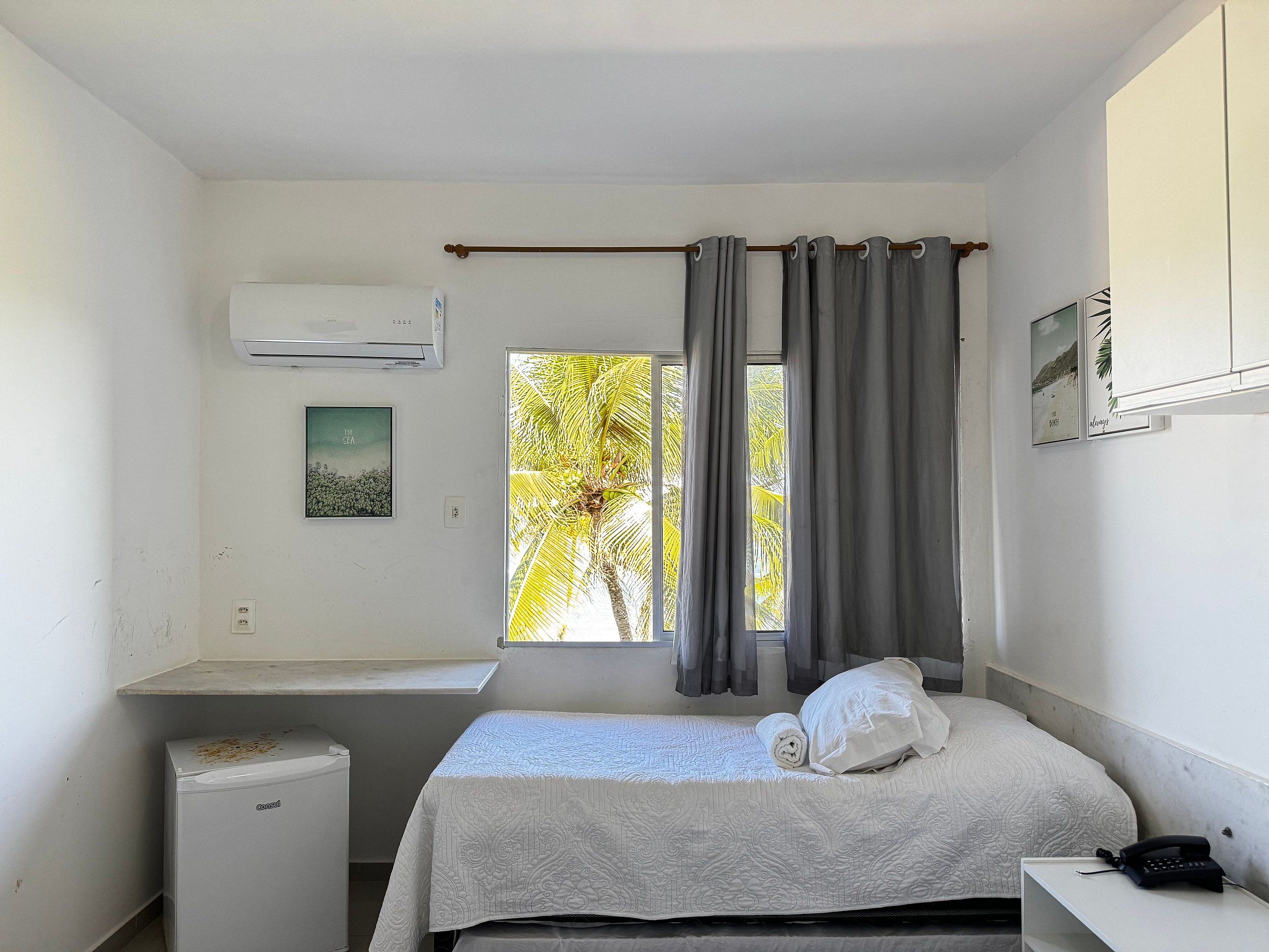 Quarto pequeno e limpo com cama, escrivaninha, ar condicionado e janela com cortinas.