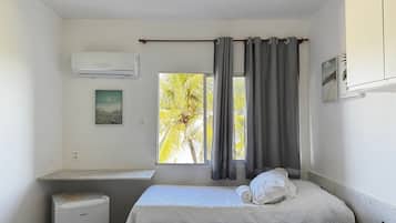 Quarto pequeno e limpo com cama, escrivaninha, ar condicionado e janela com cortinas.