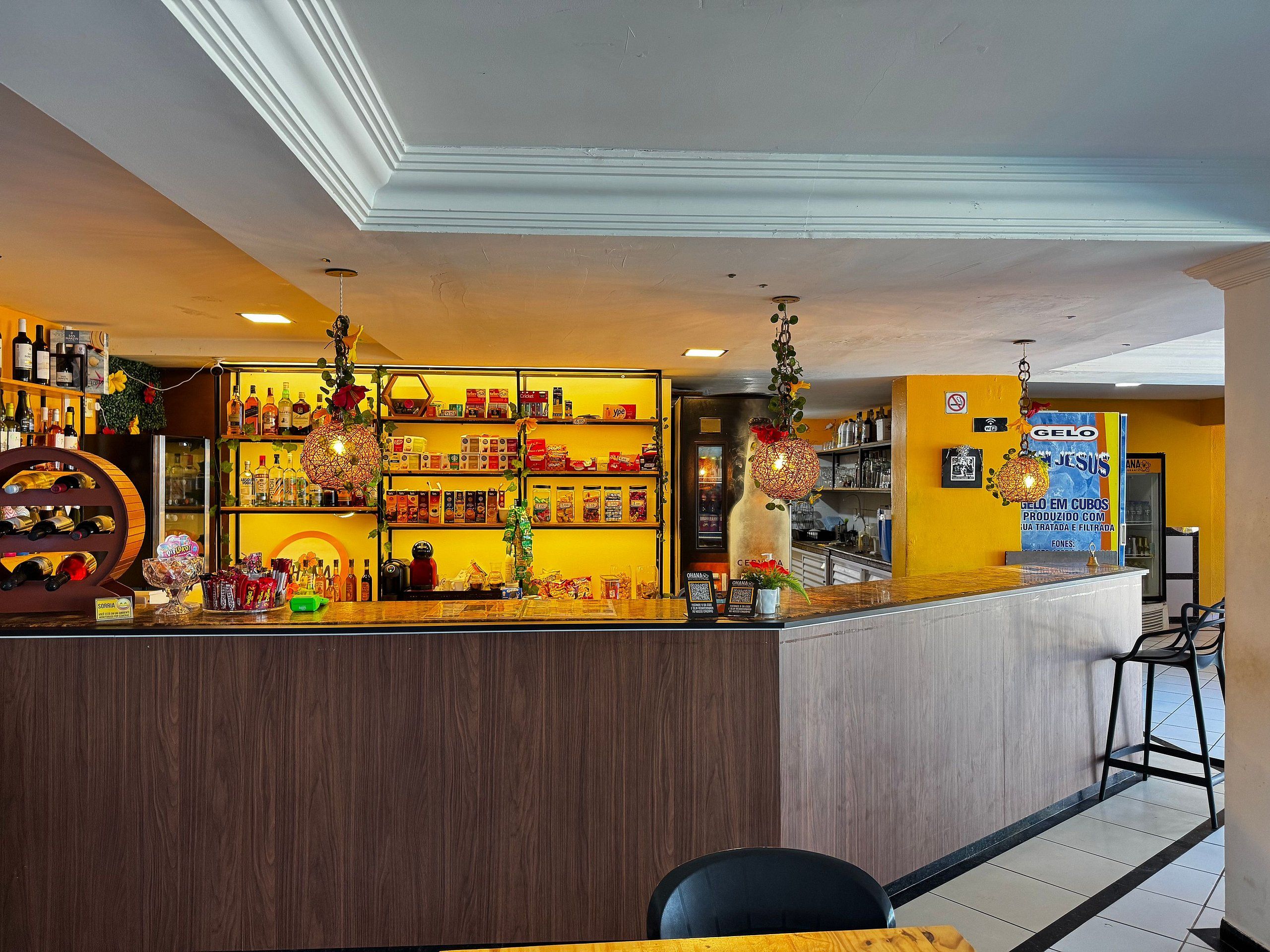 Zona de bar con estantes llenos de botellas y aperitivos, mostrador con caja registradora y luces decorativas colgantes.