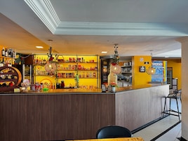 Zona de bar con estantes llenos de botellas y aperitivos, mostrador con caja registradora y luces decorativas colgantes.