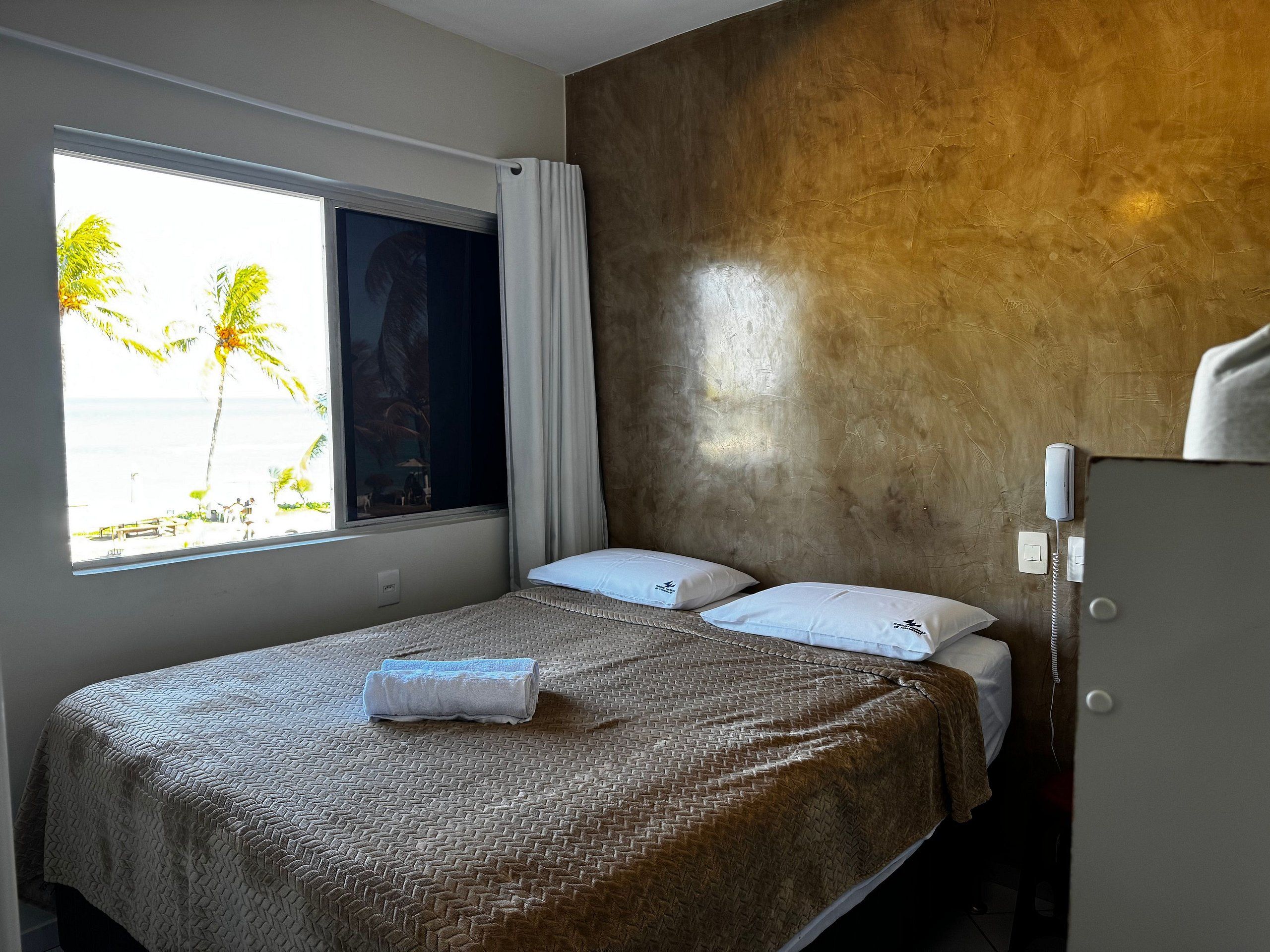 A single bed with a patterned bedspread, two pillows, a folded towel, a window with a view of palm trees, and a wooden wall.