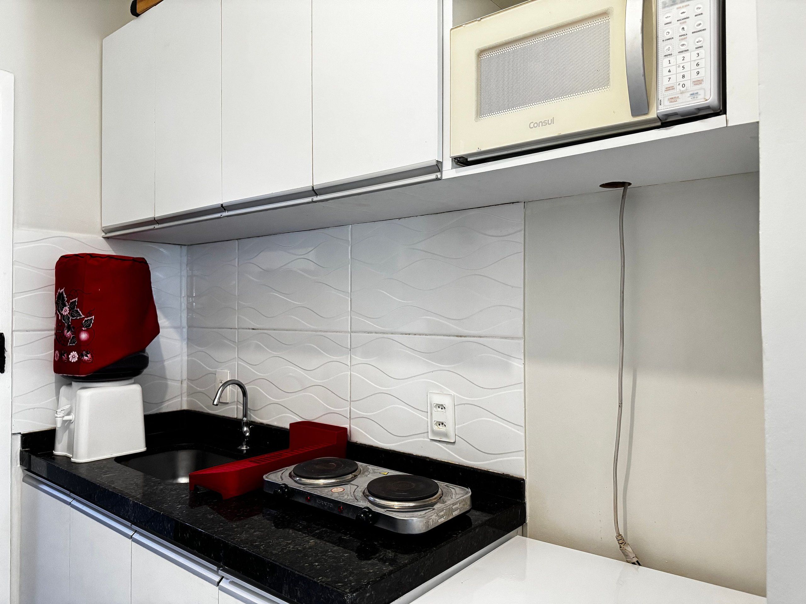 Cozinha com micro-ondas, cafeteira vermelha e fogão elétrico de duas bocas.