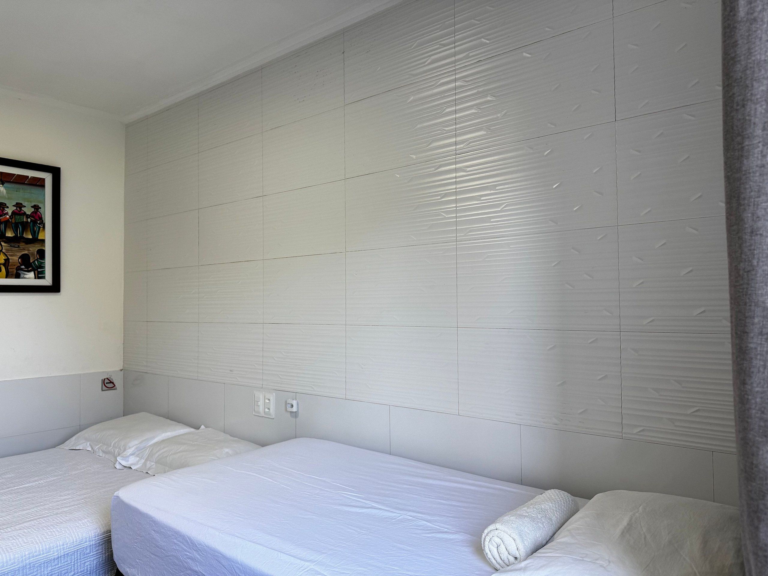 Quarto de hotel com duas camas de solteiro, cabeceira com padrão texturizado, quadro pendurado na parede e tomada.