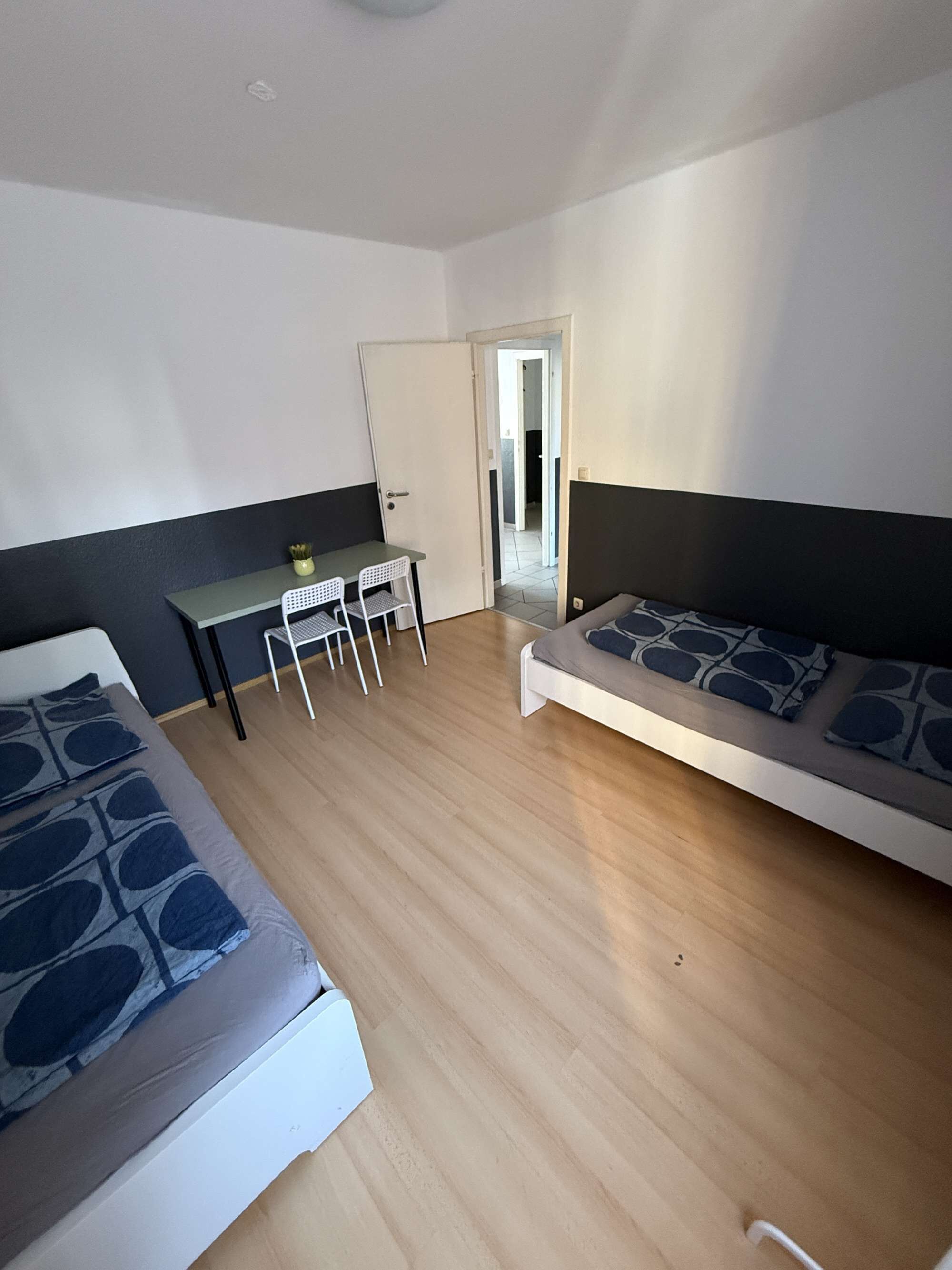 Ein Zimmer mit einem Bett, einem Schreibtisch und einem Stuhl. Eine Tür führt in einen anderen Raum.
