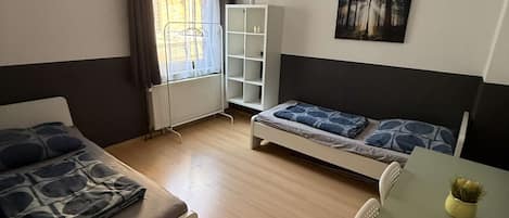 Quarto com duas camas, uma mesa, um radiador, uma janela com cortinas e uma estante de livros.