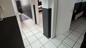 Ein Flur mit weiß gefliestem Boden, einer Tür rechts und einem Bett im Hintergrund.