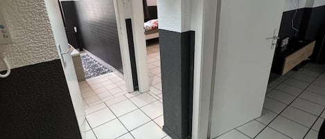 Ein Flur mit weiß gefliestem Boden, einer Tür rechts und einem Bett im Hintergrund.