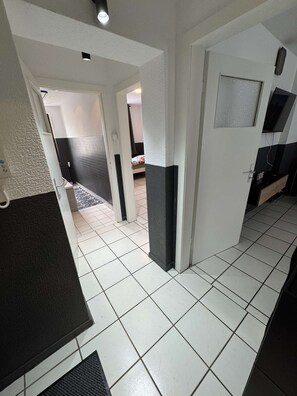 Ein Flur mit weiß gefliestem Boden, einer Tür rechts und einem Bett im Hintergrund.