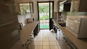 Eine moderne Küche mit Edelstahlgeräten, Fliesenboden und Blick durch eine offene Tür auf einen Außenbereich.